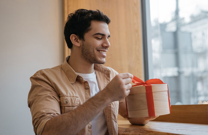 Young man happily unwrapping anniversary gift box near window, symbolizing boyfriend refuse hang anniversary gift wall.