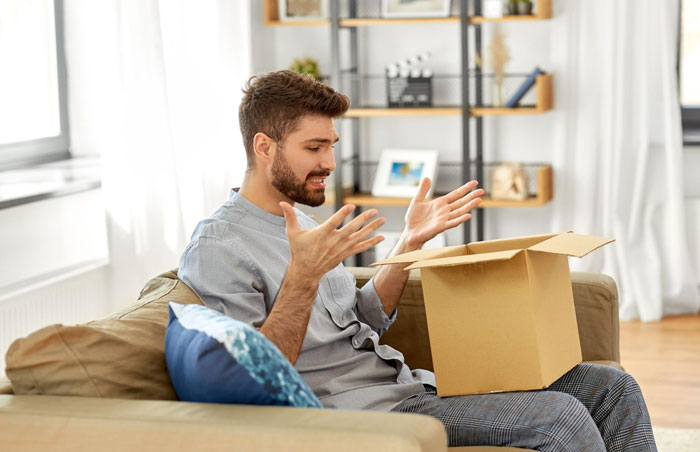 Young man on couch refusing to hang anniversary gift from wall, expressing frustration with open box on lap