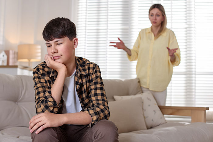 Teen stepson looks upset sitting on sofa while woman in background gestures during argument at home.