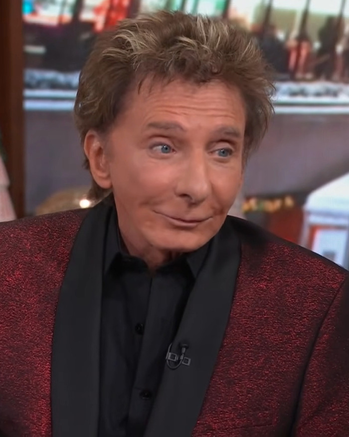 Barry Manilow wearing a red jacket and black shirt, sitting indoors with a blurred background.