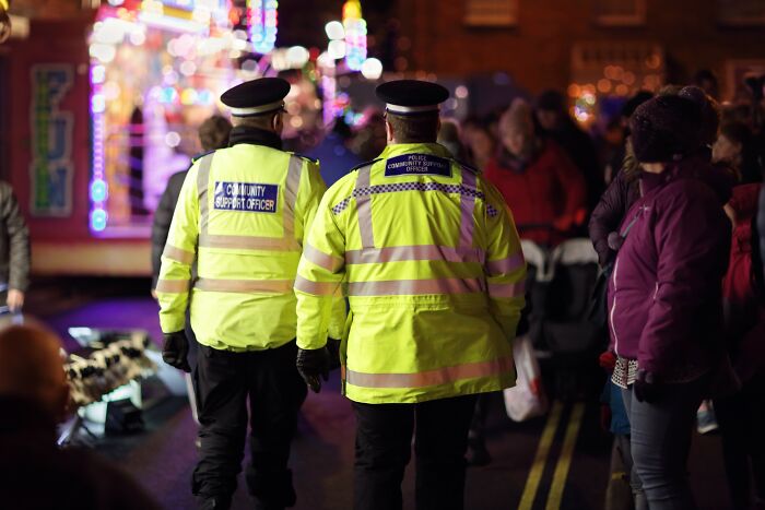 Two community support officers in high-visibility jackets walking through a crowded outdoor event at night, overseeing the area.