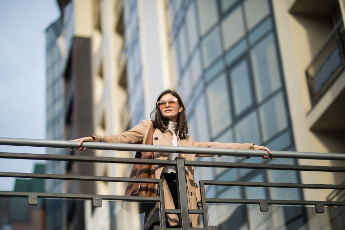 Woman wearing glasses and a beige coat standing on a balcony with modern glass buildings behind, representing solved mysteries.
