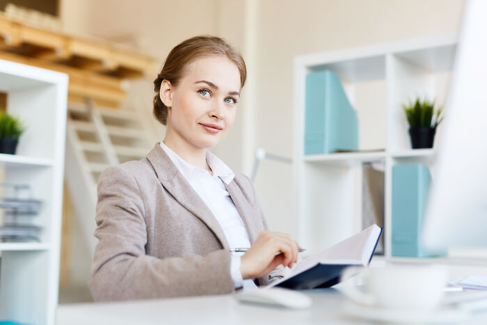 Young female accountant in a beige blazer reviewing documents at her desk, smiling confidently in a modern office setting.