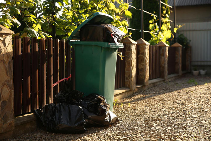Green trash bin overflowing with black garbage bags outside near a fence after neighbors’ disabled son raids the trash repeatedly Green trash bin overflowing with black garbage bags outside near a fence after neighbors’ disabled son raids the trash repeatedly