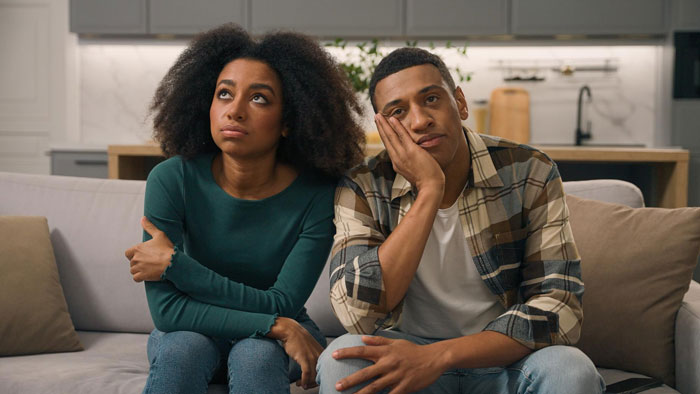 Young couple sitting on a couch looking frustrated and distant, reflecting tension over cheating accusations and family conflict.