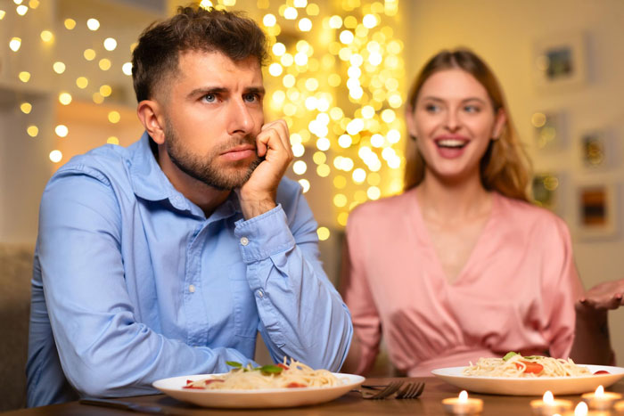 Man in blue shirt looks upset during dinner as woman in pink shirt talks, depicting conflict related to cheating accusations.