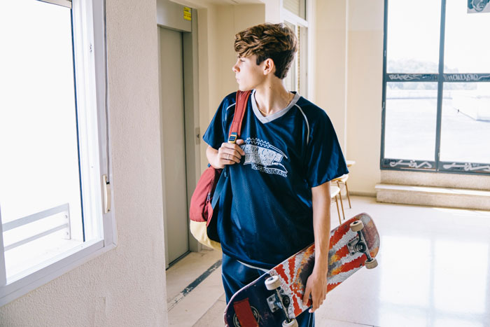 Teenage boy holding a skateboard, looking out window, highlighting care for brother with nocturnal epilepsy concerns.