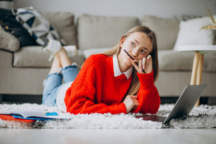 Young woman in red sweater lying on carpet, thinking with laptop open nearby, illustrating care brother nocturnal epilepsy concept.