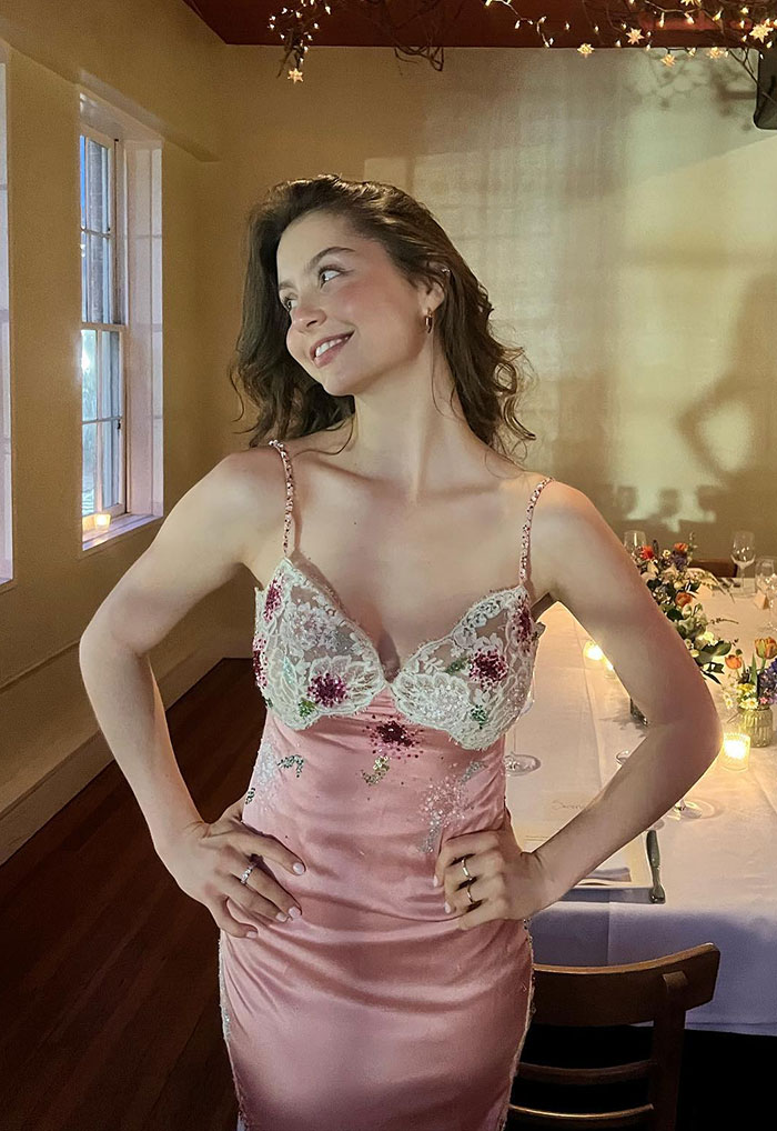 Young woman wearing a vintage pink floral dress, smiling and posing in a softly lit room at an elegant gala event.