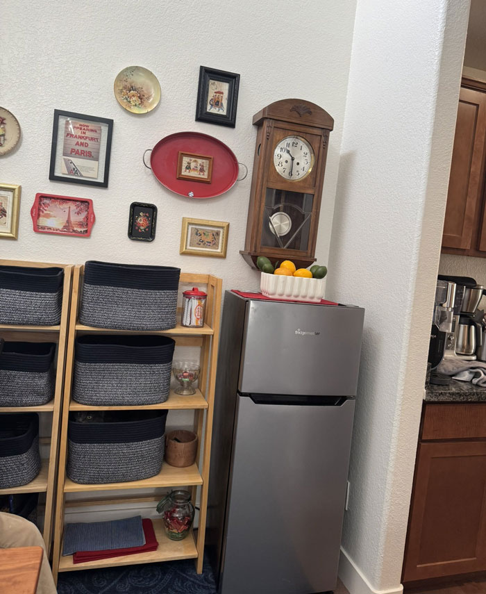 Cat blending into shelves with baskets in a cozy kitchen corner near a clock and refrigerator.