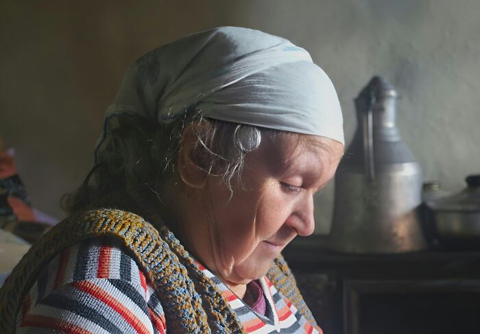 Elderly woman wearing a headscarf, sitting thoughtfully in a dimly lit room, hinting at dark secrets and hidden stories.