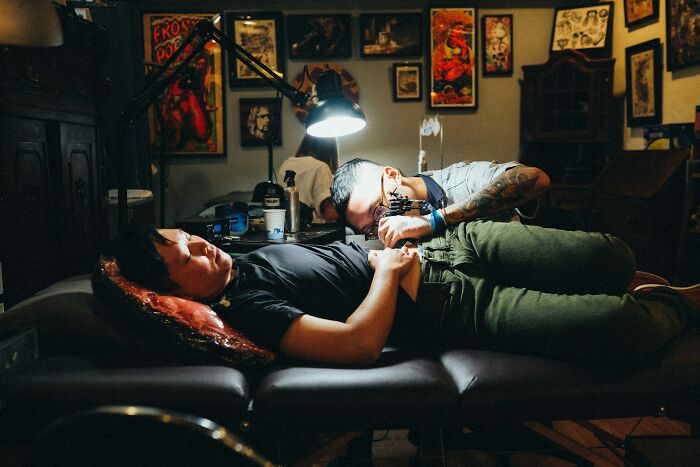 Tattoo artist working on a client in a dimly lit studio filled with framed artwork on the walls.