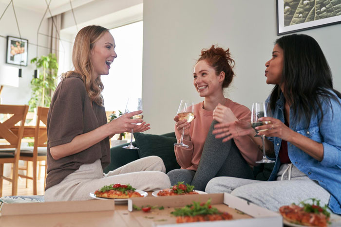 Three women enjoying pizza and wine while engaged in a lively conversation during a friend visit abroad.