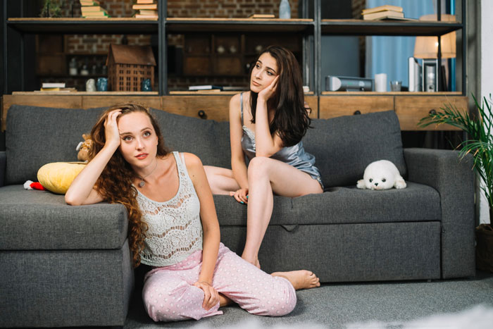 Two women in pajamas looking concerned on a couch, reflecting on a friend&rsquo;s intoxicated partner serenading her in bed.