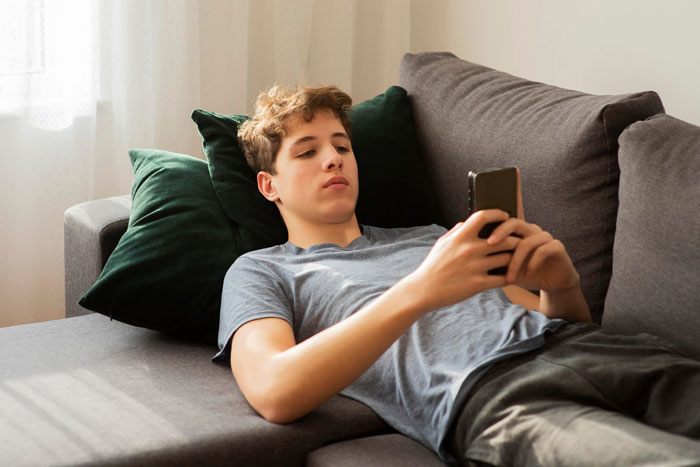 Young man mooching off cousin lounging on couch and using smartphone, avoiding paying $400 rent demand.