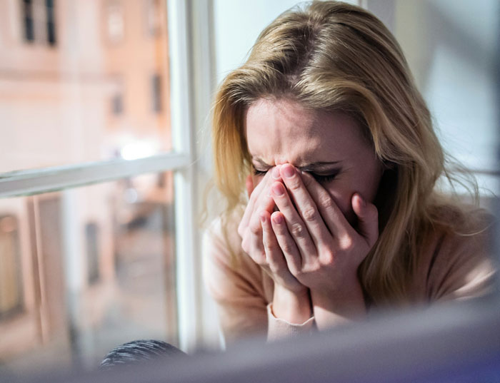 Woman covering her face in distress by a window, illustrating emotional pain from a baby lie and relationship deception.