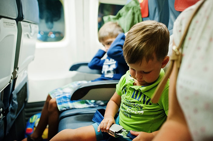 Young disruptive kid on a plane adjusting seatbelt with upset mom and another child in the background