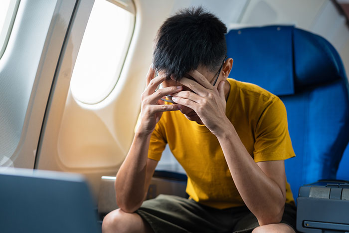 Man on plane covering face in frustration while sitting next to window, upset after telling disruptive kid to shut up.