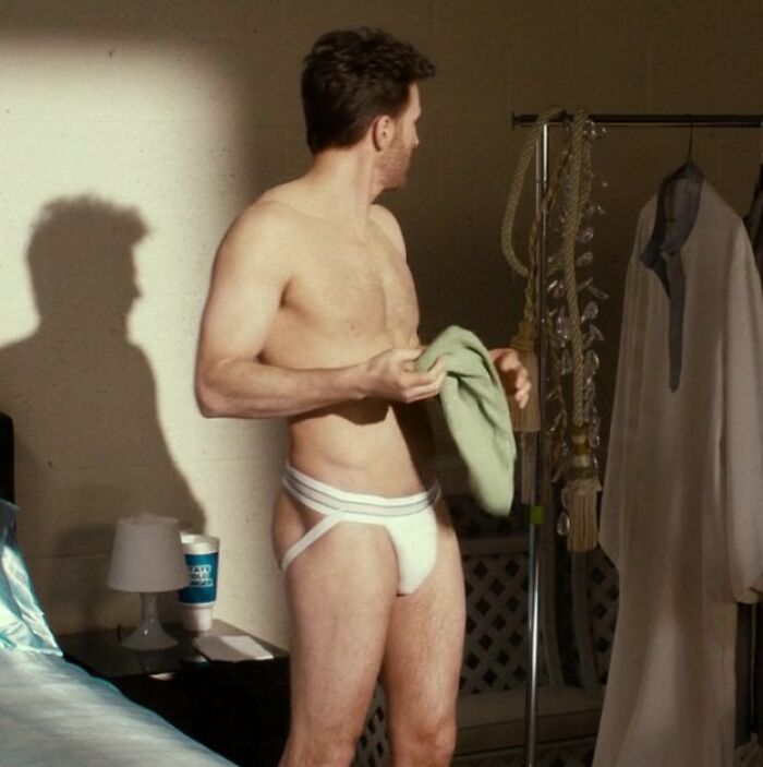 Chris Evans wearing a white jock strap, standing shirtless in a room, holding a green cloth with clothing rack nearby.