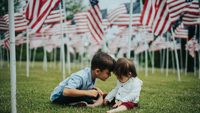 Two children sitting on grass among American flags, symbolizing hidden dark secrets that could ruin people’s lives.