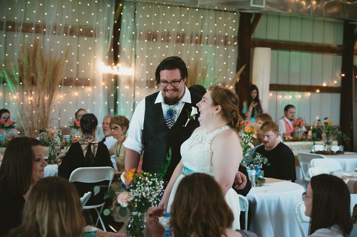 Bride and groom smiling and interacting with guests at a decorated wedding reception with string lights and floral arrangements.