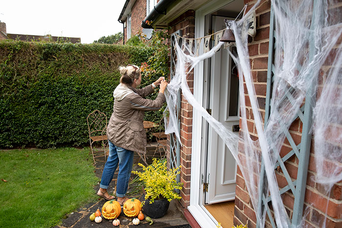 Woman decorating house with Halloween decor including spider webs and jack-o-lanterns, facing neighbor threats over Halloween decorations