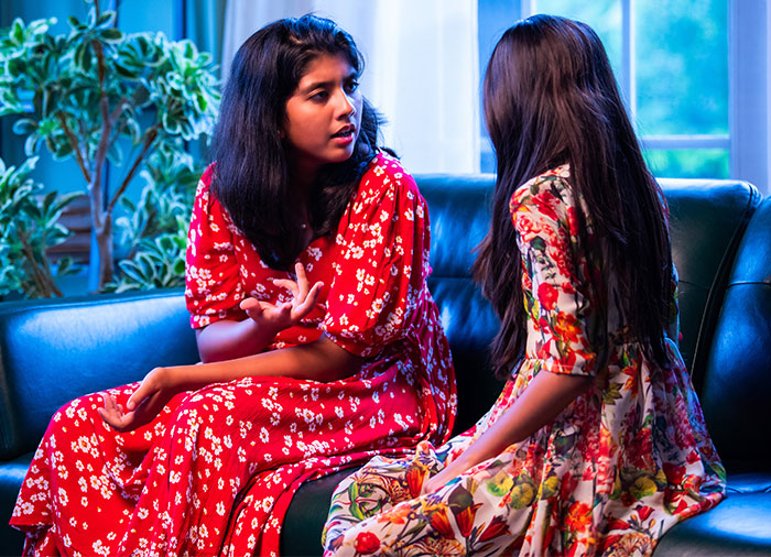 Two women in floral dresses having a serious conversation inside a living room, highlighting babysitting struggles.