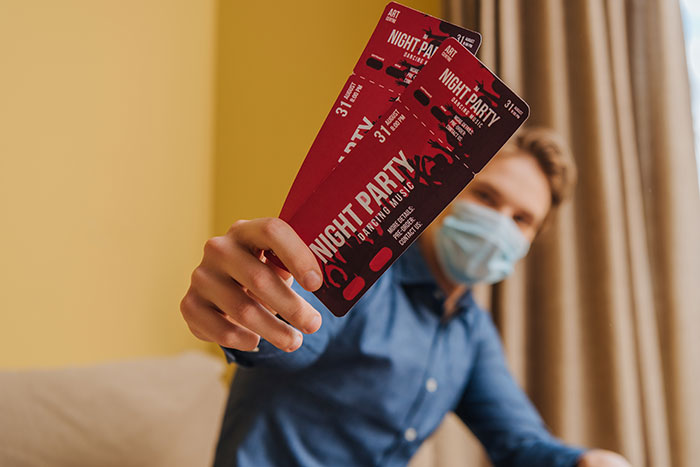 Person wearing mask holding two night party tickets indoors, illustrating coworker show scam realization.