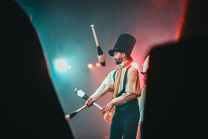 Male performer in a tall hat juggling clubs on stage during a show with dramatic lighting and an audience silhouette.