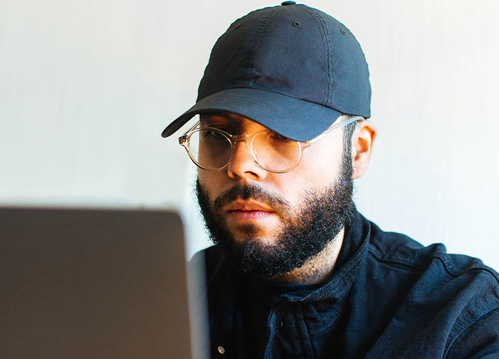 Man with glasses and cap focused on laptop screen in a casual setting, related to coworker wearing makeup discussion.