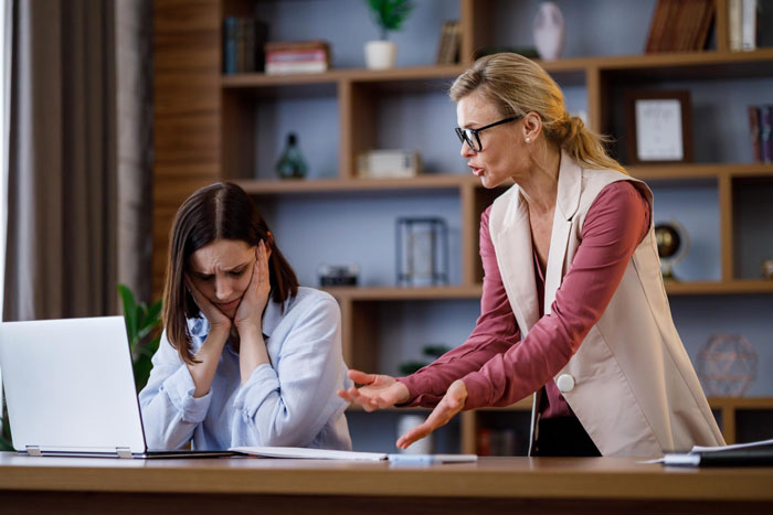 Two coworkers in an office, one upset and holding her head, the other angrily gesturing during a heated argument.