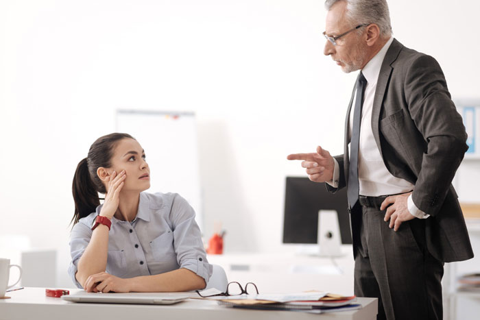 An older man in a suit reprimanding a young female employee at her desk, illustrating an entitled employee conflict.