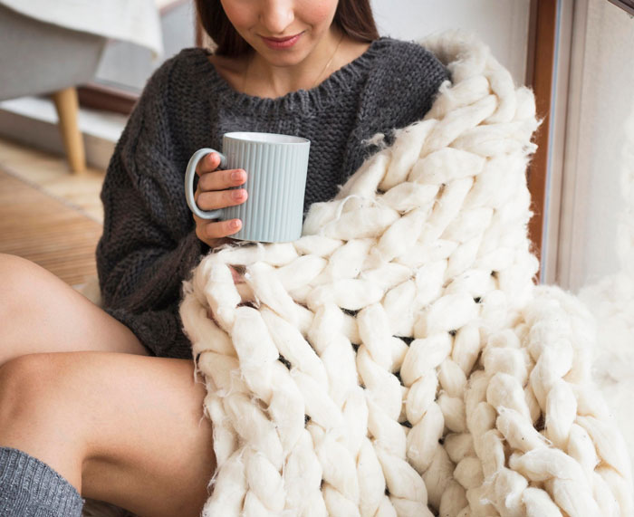 Woman wrapped in a huge chunky knit blanket holding a cup, representing knitting hobby and cozy crafting indoors.