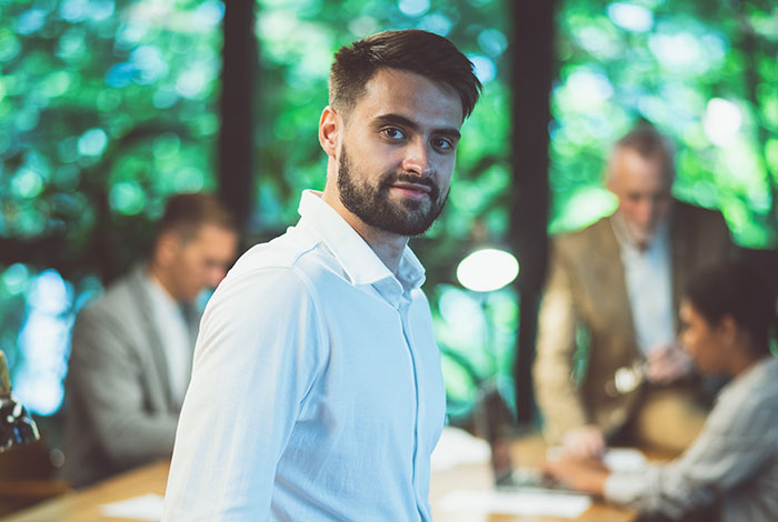Young man in white shirt looking at camera in office setting with coworkers discussing work nepotism in background.