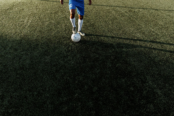 Soccer player in blue jersey controlling ball on dark grass field with shadow, representing spooky urban legends theme.
