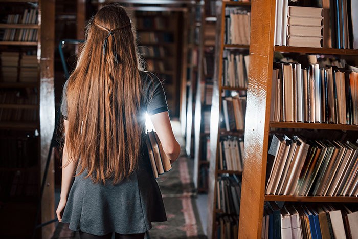Young woman with long hair holding books, exploring a dimly lit library, evoking spooky urban legends atmosphere.