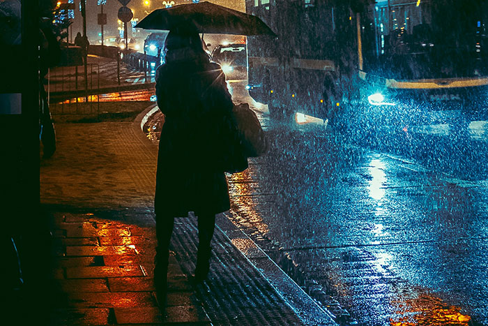 Person walking with umbrella on rainy city street at night, evoking spooky urban legends atmosphere.