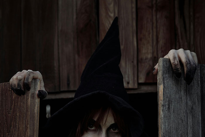 Person in a dark witch hat with pale hands emerging from behind old wooden doors for spooky urban legends theme