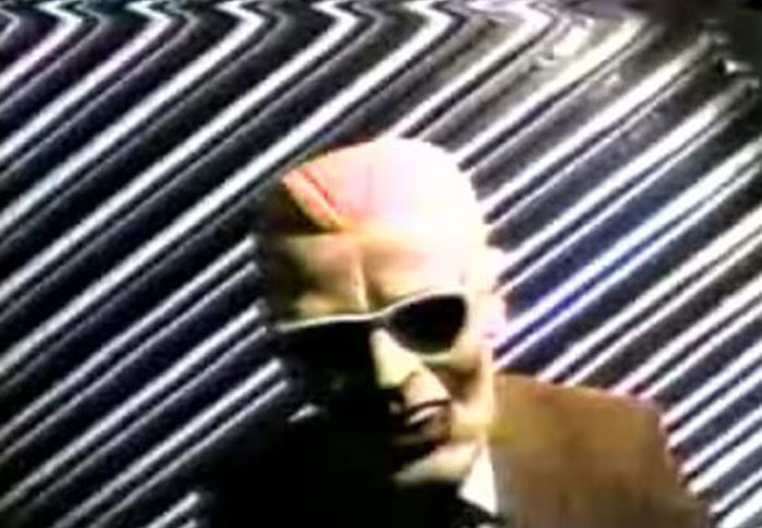 Creepy found footage video frame showing a distorted figure with sunglasses against a static background.