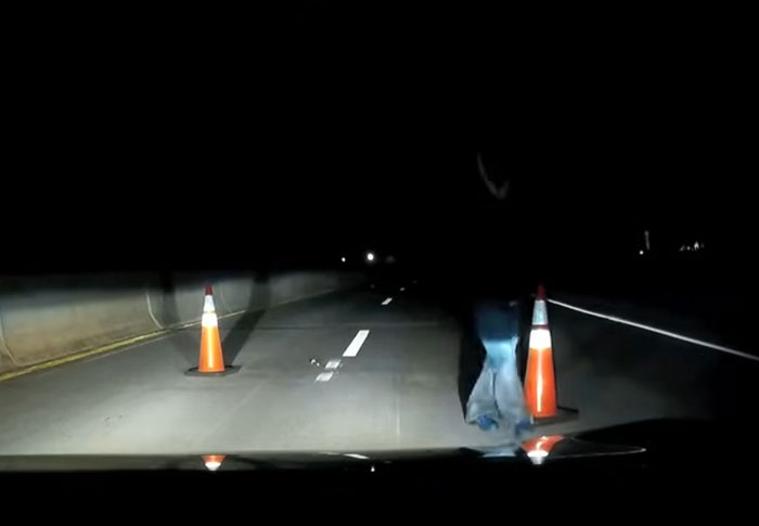 A dark, eerie night scene captured in creepy found footage with traffic cones and a shadowy figure on the road.
