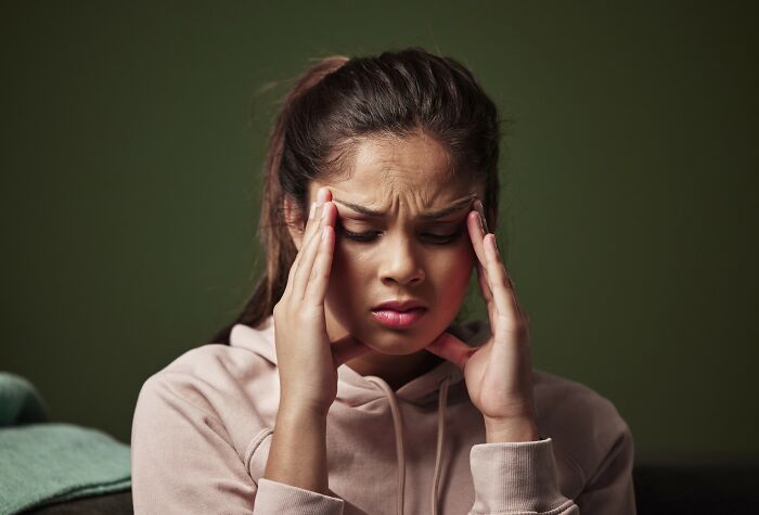 Young woman in a pink hoodie showing signs of medical problems, holding her head in pain with a distressed expression.