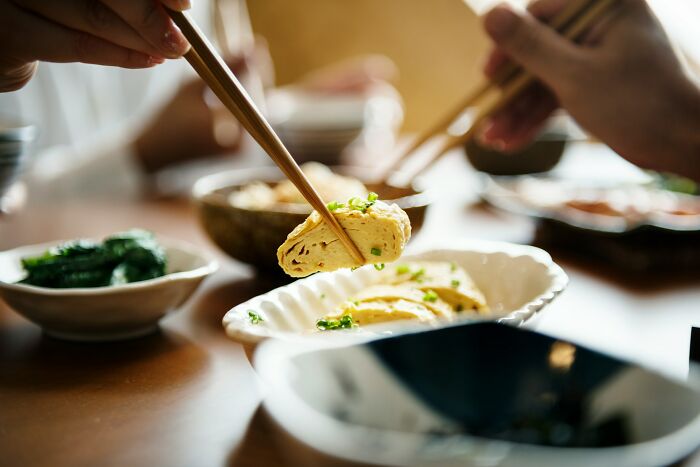 Hands using chopsticks to pick up a rolled omelette from a white dish during a casual meal setting.