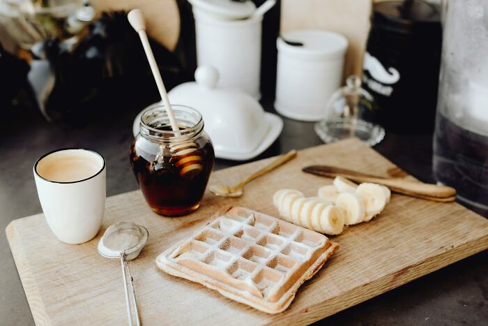 Waffle and sliced bananas on a wooden board with honey jar and cup of coffee in a cozy kitchen setting.