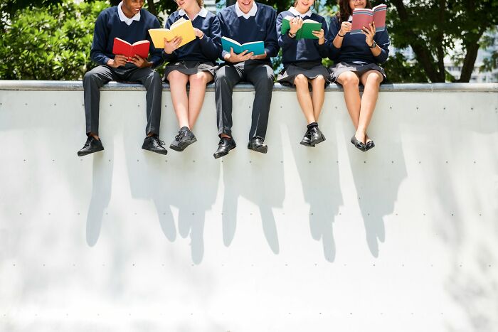 Group of popular kids in school uniforms sitting on a wall reading books, showing shocking school behaviors that caused outcasts.