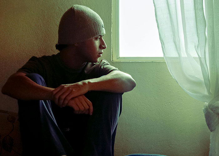 Teen sitting by a window in a dim room, appearing troubled and torn about seeing biological mom and feeling second-rate. Teen sitting by a window in a dim room, appearing troubled and torn about seeing biological mom and feeling second-rate.