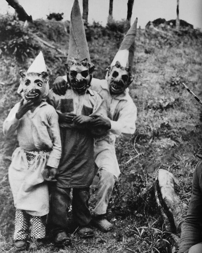 Three children in vintage Halloween costumes with creepy masks and pointed hats posing outdoors on a grassy hill.