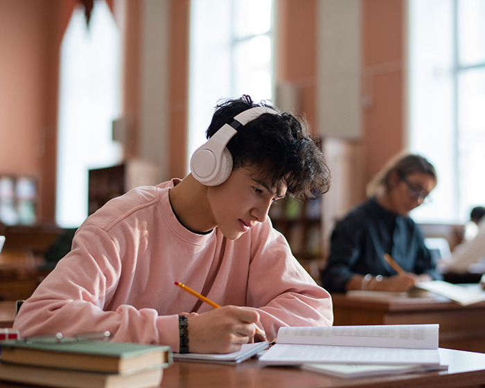 Teen student wearing headphones studying intently with books and notes in a quiet classroom setting involving teacher interaction.