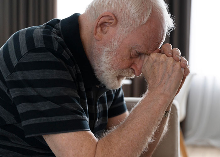 Elderly dad upset about daughter&rsquo;s home, leaning forward with hands clasped, showing worry and stress indoors.
