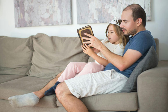 Man reading a book with his young daughter on the couch, highlighting challenges dating men with kids again.