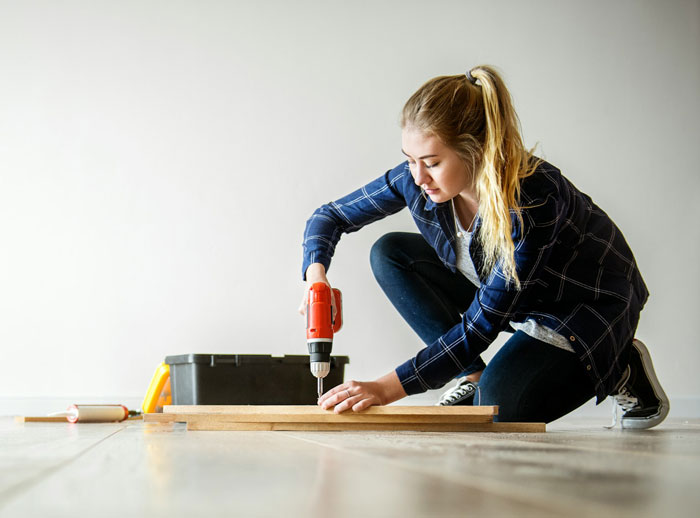 Young woman using a power drill on wooden board, symbolizing a daughter involved in a room remodeling project.